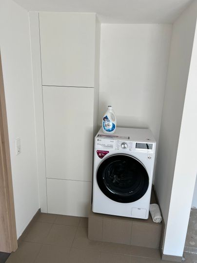 Eine moderne Waschmaschine steht in einem hellen Raum. Neben der Maschine befindet sich ein Flasche Waschmittel.
