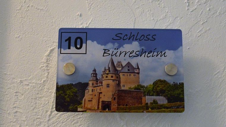 Ein Schild mit der Aufschrift „Schloss Bürresheim“ und der Nummer 10. Das Bild zeigt ein schönes Schloss umgeben von Bäumen und einem blauen Himmel.