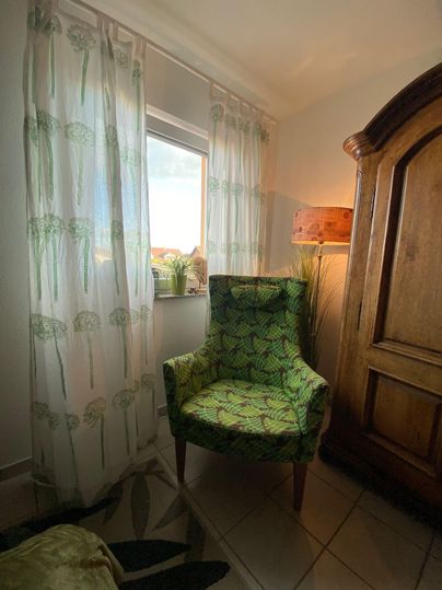 Ein gemütlicher Sessel mit grünem Muster steht in einer Ecke. Durch das Fenster fällt sanftes Licht und die Vorhänge haben ein pflanzenartiges Design.