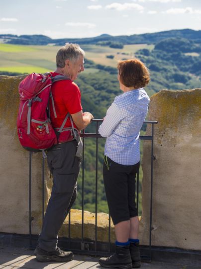 Ein Paar betrachtet die Landschaft von einer Aussichtsplattform. Im Hintergrund sind grüne Hügel und ein blauer Himmel zu sehen.