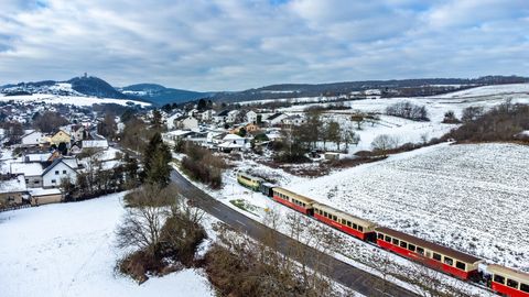 Eine bunte Lokomotive steht im Schnee, während Menschen auf dem Bahnsteig spazieren. Die Landschaft ist winterlich und malerisch mit schneebedeckten Bäumen.