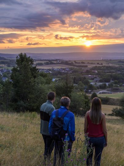 Drei Personen stehen auf einer Wiese und betrachten den Sonnenuntergang über der Landschaft. Der Himmel ist bunt und die Atmosphäre ist friedlich.