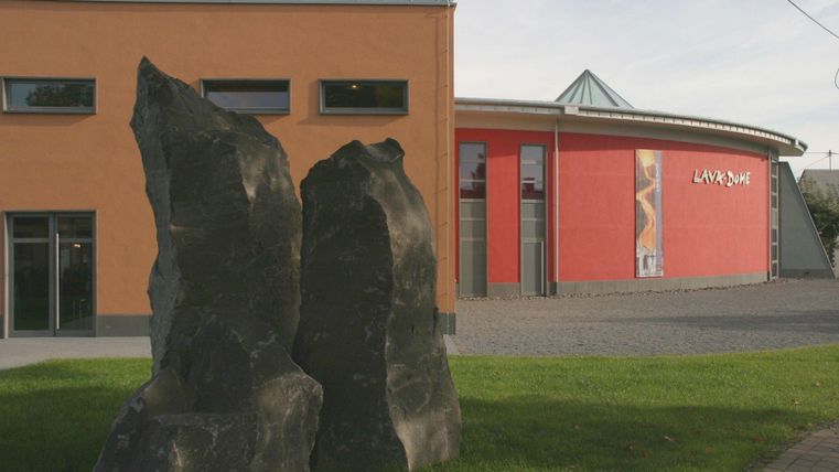 Ein modernes Gebäude mit orangefarbener Fassade und Rundung. Im Vordergrund stehen zwei große Steinskulpturen auf einer grünen Wiese.