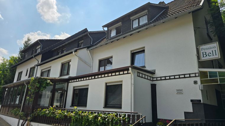 Ein Gästehaus mit zwei Etagen und einem einladenden Eingangsbereich. Die Fassade ist hell und von grünen Pflanzen umgeben.