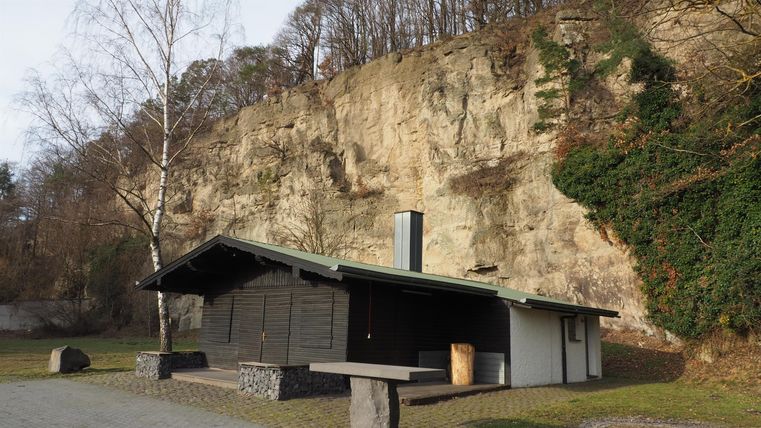 Ein gemütliches Holzhaus steht vor einer hohen, bewaldeten Felswand. Die Umgebung ist ruhig und natürlichen.