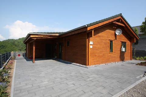 Ein modernes Holzhaus mit einer großzügigen Terrasse aus Pflastersteinen. Es liegt in einer grünen Umgebung unter einem klaren blauen Himmel.