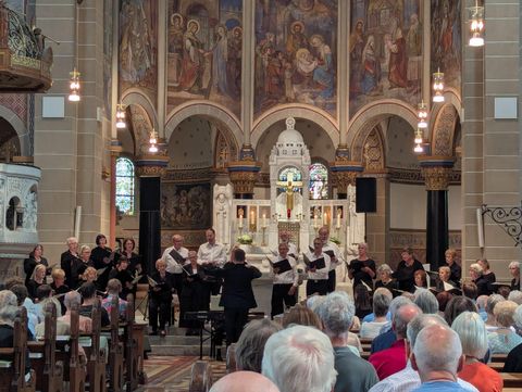 Een koor staat in een kerk en geeft een concert. Veel toeschouwers zitten in het publiek en genieten van de uitvoering.