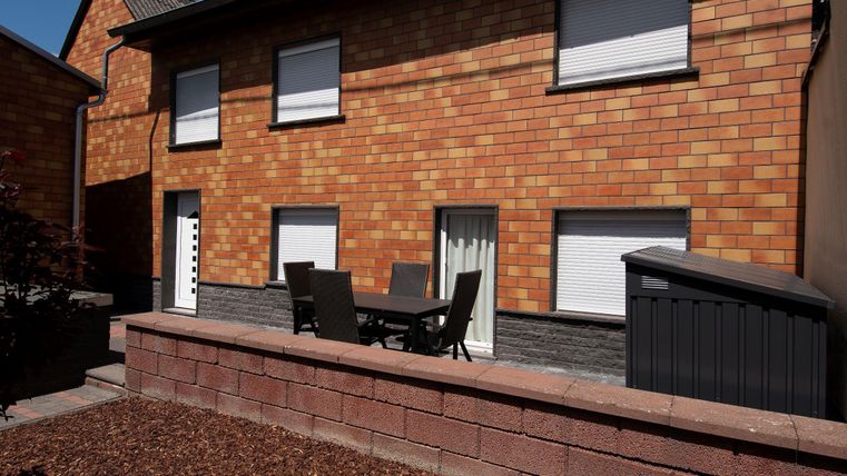 Ein modernes, zweigeschössiges Haus mit Ziegelfassade. Im Vordergrund steht ein gemütlicher Gartentisch mit Stühlen.