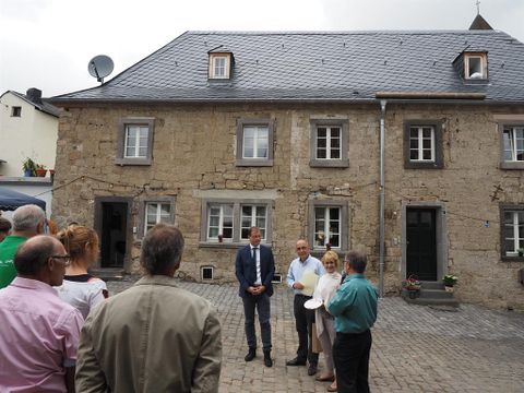 Eine Gruppe von Menschen steht vor einem alten Steinhaus mit grauem Dach. Einige Personen sprechen miteinander, während andere aufmerksam zuhören.