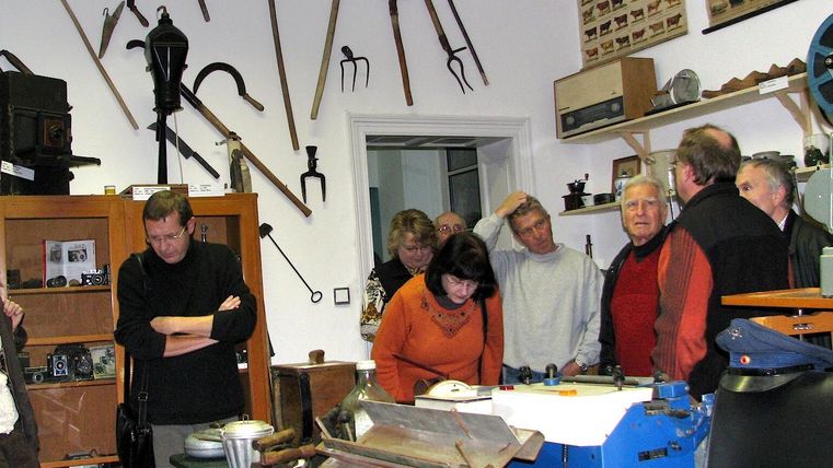 Eine Gruppe von Menschen steht in einem Museum voller alter Werkzeuge und Geräte. An den Wänden hängen verschiedene Werkzeuge und Objekte, die die Besucher interessieren.