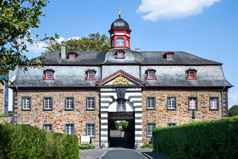 Ein historisches Gebäude mit roten und grauen Akzenten. Es hat eine zentrale Toröffnung und ist von grünen Hecken umgeben.