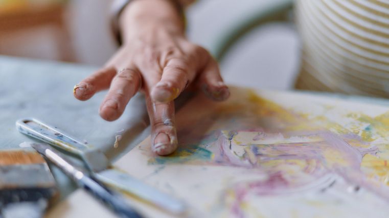 Eine Hand, die mit Farben auf einem Blatt Papier arbeitet, ist im Vordergrund zu sehen. Pinsel und Farbspuren deuten auf kreative Aktivitäten hin.