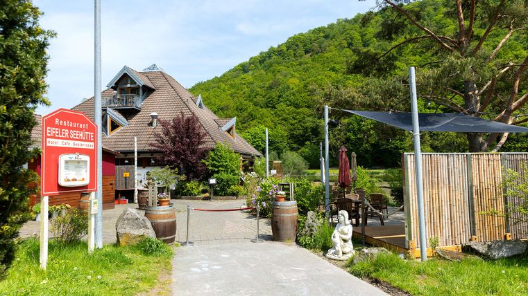 An inviting restaurant with a beautiful entrance area, surrounded by trees and greenery. In the background, gentle hills are visible, creating a tranquil atmosphere.