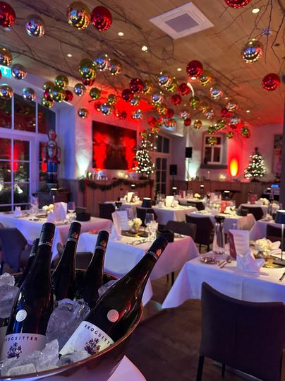 Ein festlich dekorierter Saal mit Tischen, die für eine Feier gedeckt sind. Über dem Raum hängen bunte Weihnachtskugeln und der Raum ist sanft beleuchtet.