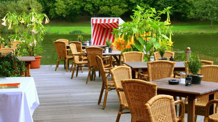 Eine gemütliche Terrasse am Wasser mit Holztischen und Rattan-Stühlen. Umgeben von grünen Pflanzen und einem rot-weiß gestreiften Sonnenschirm.