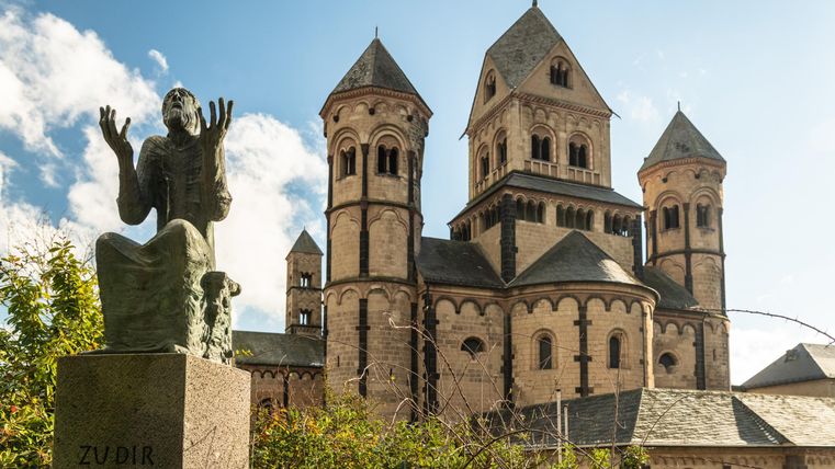 Eine Statue eines Betenden steht vor einer beeindruckenden Kirche mit Türmen. Der Himmel ist blau und es gibt einige Wolken.