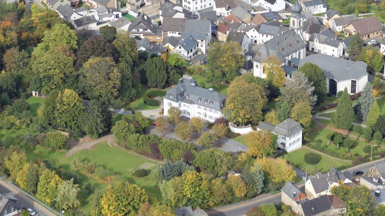 An aerial view of a city with many buildings and lush greenery. In the foreground, there is a large garden with trees and a white villa.