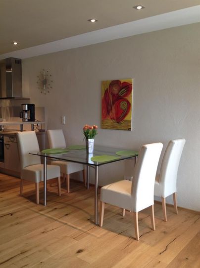 Ein moderner Essbereich mit einem Glastisch und vier weißen Stühlen. An der Wand hängt ein farbenfrohes Gemälde und frische Blumen stehen in einer Vase auf dem Tisch.