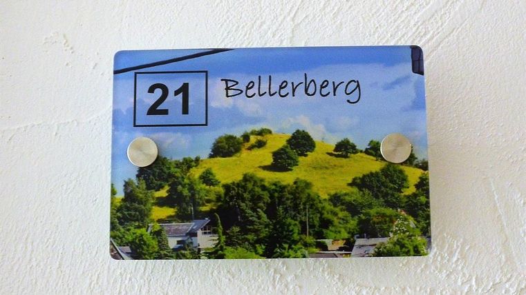 Eine Haustürnummer mit der Aufschrift "21 Bellerberg" wird auf einer blauen Tafel angezeigt. Im Hintergrund sind grüne Hügel und Bäume zu sehen.