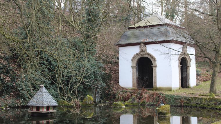 Ein kleines, historisches Gebäude steht neben einem Teich, umgeben von Bäumen. Im Hintergrund ist eine bewaldete Landschaft sichtbar.