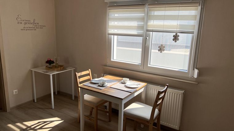 Ein heller, gemütlicher Essbereich mit einem kleinen Tisch und zwei Stühlen. Neben dem Tisch befindet sich ein Fenster mit Jalousien, das für viel Tageslicht sorgt.