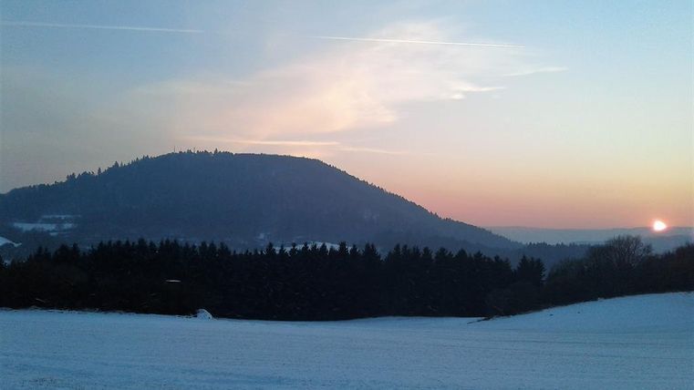 Eine winterliche Landschaft mit schneebedecktem Feld und einem Hügel im Hintergrund. Der Sonnenuntergang färbt den Himmel in sanften Farbtönen.