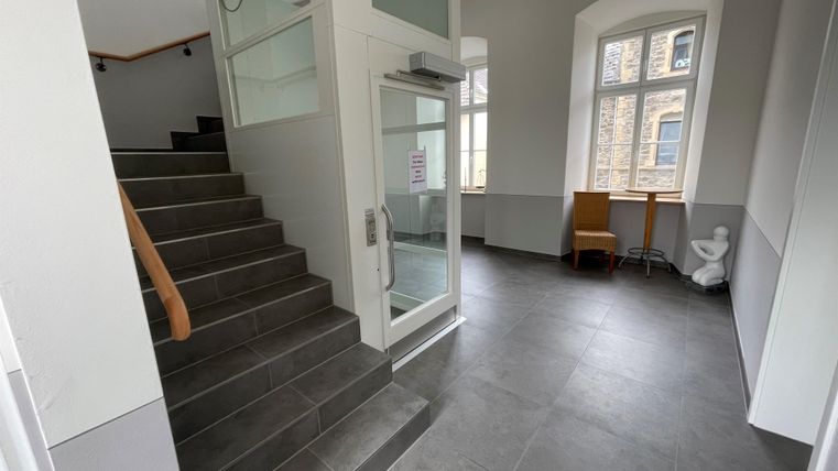 Ein moderner Eingangsbereich mit einer Treppe und großen Fenstern. Es gibt einen kleinen Tisch und einen Stuhl in der Ecke.