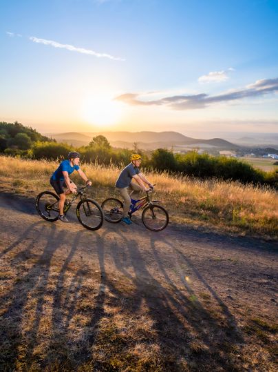 Zwei Fahrradfahrer fahren einen Radweg entlang. im Hintergrund sieht man ein abgeerntetes Feld und einen Sonnenuntergang.