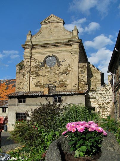Historisches Gebäude mit verwitterter Fassade und rundem Fenster, umgeben von Pflanzen und Blumen.
