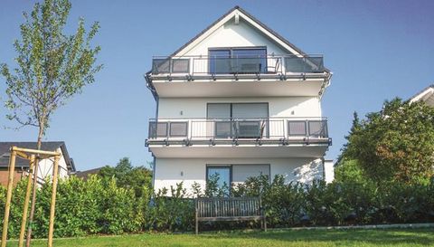Ein modernes Haus mit mehreren Balkonen und großen Fenstern. Vor dem Haus befindet sich eine Grünfläche mit einer Bank und einem kleinen Baum.