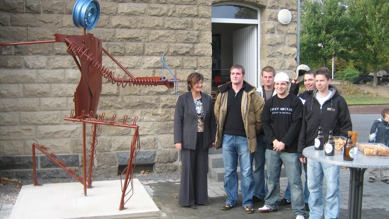 Eine Gruppe von Personen steht vor einem Gebäude mit einer Metallskulptur. Die Skulptur zeigt eine humanoide Figur mit einem Rad als Kopf.