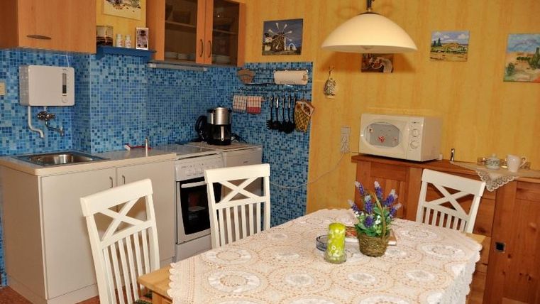 Eine helle Küche mit blauen Fliesen und einem Esstisch aus Holz. Der Tisch ist mit einer Tischdecke dekoriert und es gibt einen Mikrowelle sowie einen Wasserkocher.