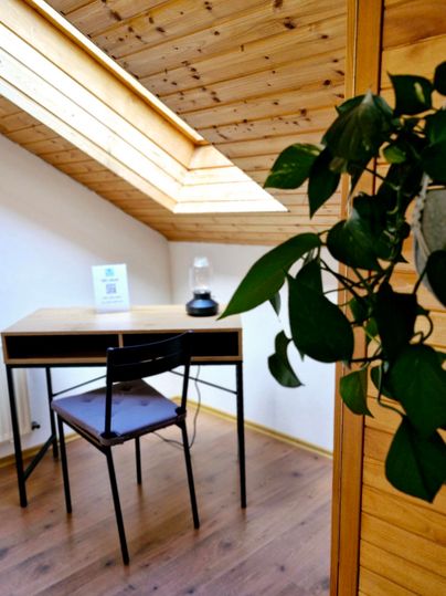 Ein heller Raum mit einem Holztisch und einem Stuhl unter einem Dachfenster. Im Hintergrund ist eine Topfpflanze zu sehen.