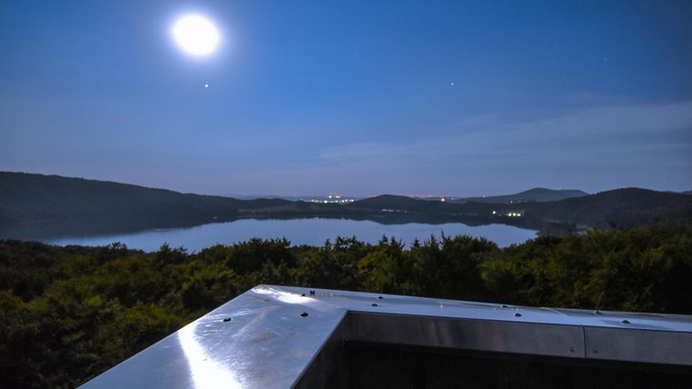 Eine ruhige Nachtansicht mit einem klaren Mond über einem stillen See. Im Hintergrund sind sanfte Hügel und das sanfte Licht der Stadt zu sehen.