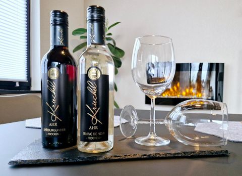 Two bottles of wine, one red and one white, are standing on a black coaster. Next to them is an empty wine glass and an upside-down glass, while a warm fire is burning in the background.