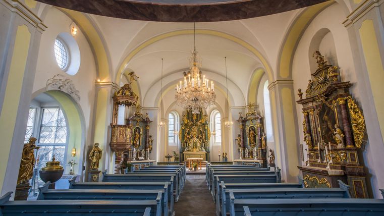 A beautiful church with a bright interior and elaborate statues. The benches are blue and the chandelier provides pleasant light.