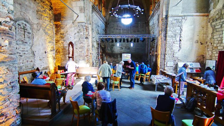 Ein historischer Raum mit steinernen Wänden, der als Café genutzt wird. Menschen sitzen an Tischen und genießen ihre Zeit in dieser gemütlichen Atmosphäre.