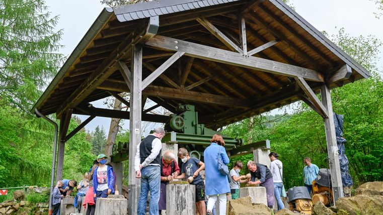 Eine Gruppe von Menschen arbeitet unter einem Holzpavillon. Im Hintergrund sieht man eine alte Maschine und viel grüne Umgebung.