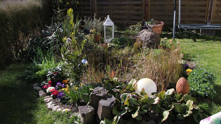 Ein schöner Garten mit bunten Blumen, Grünpflanzen und dekorativen Elementen. Im Hintergrund steht ein Holzzaun.