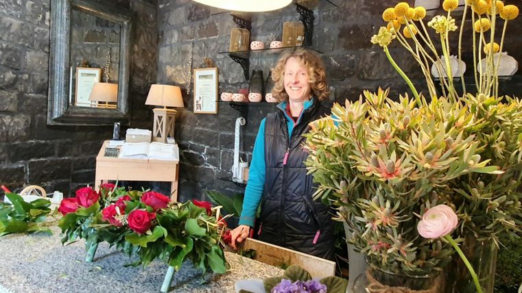 A blooming flower boutique with a friendly saleswoman. Colorful flower arrangements are on the table and the walls are made of stone.
