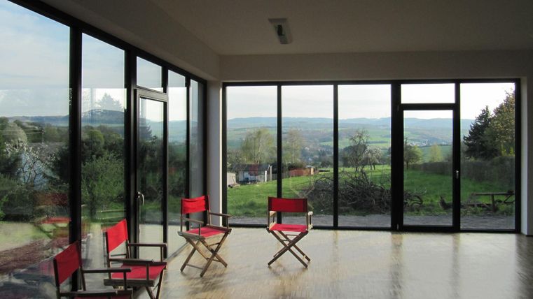 Ein moderner Raum mit großen Fenstern und Blick auf die grüne Landschaft. Rote Stühle stehen im Vordergrund.