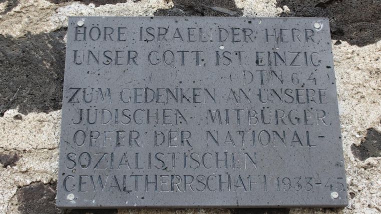 Eine Gedenktafel mit dem Text "Höre Israel, der Herr unser Gott ist ein einziger" und einer Inschrift zu Ehren der jüdischen Mitbürger, die unter der nationalsozialistischen Gewaltherrschaft litten. Die Tafel ist aus Metall und befindet sich an einer Wand.