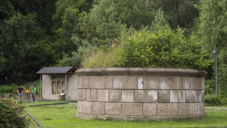 Eine runde, steinerne Struktur mit Vegetation oben, umgeben von einer grünen Wiese. Im Hintergrund ist ein einfaches Gebäude und einige Spaziergänger zu sehen.