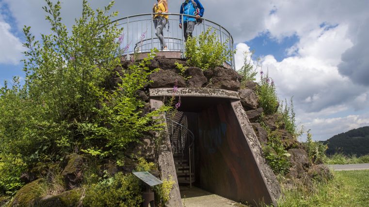 A stone observation platform with two people on it. Surrounded by green plants and a bright, cloudy sky.