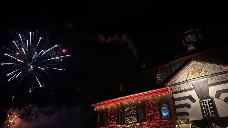 Ein festlich beleuchtetes Gebäude bei Nacht mit buntem Feuerwerk im Hintergrund. Die Szenerie strahlt eine feierliche Stimmung aus.