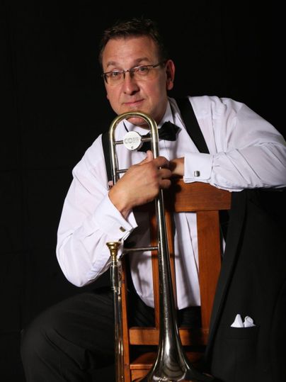 Een man in formele kleding zit op een stoel en houdt een trombone vast. De achtergrond is zwart, wat de nadruk legt op de muzikant en zijn instrument.