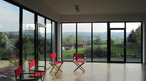 Ein heller Raum mit großen Fenstern und Blick auf eine grüne Landschaft. Im Raum stehen drei rote Stühle.