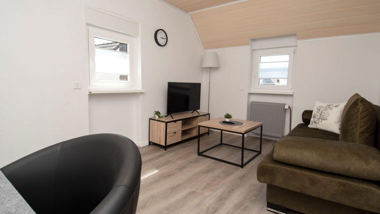 Ein modernes Wohnzimmer mit einem gemütlichen Sofa und einem Fernseher. Große Fenster sorgen für viel Tageslicht.