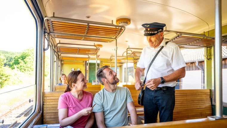 Eine Person in Uniform spricht mit zwei Fahrgästen in einem Zug. Die Szene zeigt eine freundliche Interaktion in einem modernen, hellen Zugabteil.