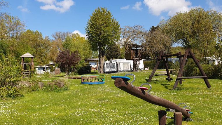 Ein weitläufiger Spielplatz mit Rasen, Spielgeräten und Bäumen. Im Hintergrund sind Wohnwagen und ein klarer Himmel zu sehen.